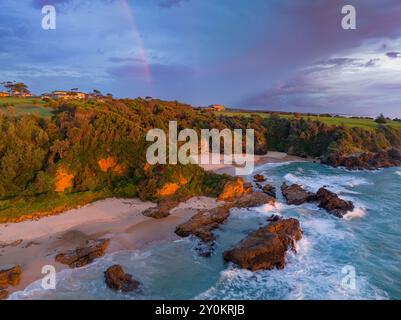 Aus der Vogelperspektive sehen Sie dramatische Beleuchtung und einen schwachen Regenbogen über einer grünen Landzunge und der zerklüfteten Küste von Narooma an der Südküste von New South Wales Stockfoto