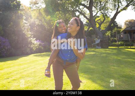 Die Tochter mit dem Huckepack fahren, die Mutter lächelt und das Sonnenlicht im Garten genießt Stockfoto