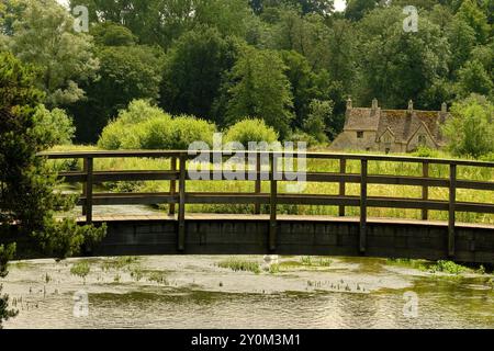 Blick auf eine hölzerne Fußgängerbrücke über den Fluss Coln zur Rack Isle Wasserwiese und den Cottages im Cotswold Dorf Bibury, England, Großbritannien Stockfoto