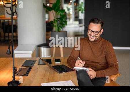 Hochwinkelansicht des lächelnden Geschäftsmannes, der Notizen schreibt, während er am Schreibtisch im Büro über dem Laptop arbeitet Stockfoto