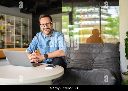 Porträt eines lächelnden männlichen Freelancers mit gefalteten Händen, sitzend mit Laptop auf Sofa in einem modernen Büro Stockfoto