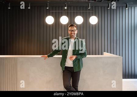 Porträt des Rezeptionisten in Brille, der in die Kamera lächelt und am Lobbyschreibtisch im beleuchteten Hotel steht Stockfoto