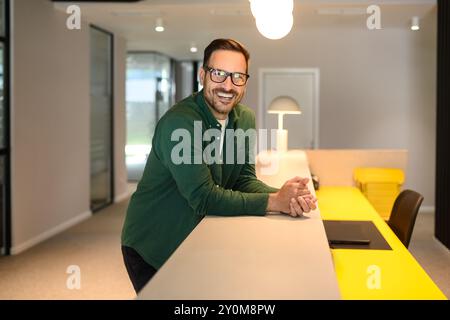 Porträt eines glücklichen männlichen Managers mit gefalteten Händen und in Brille, der an der beleuchteten Rezeption steht Stockfoto