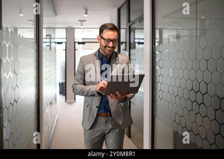Porträt eines Entwicklers, der lächelt und online über Laptop recherchiert, während er im Bürokorridor läuft Stockfoto