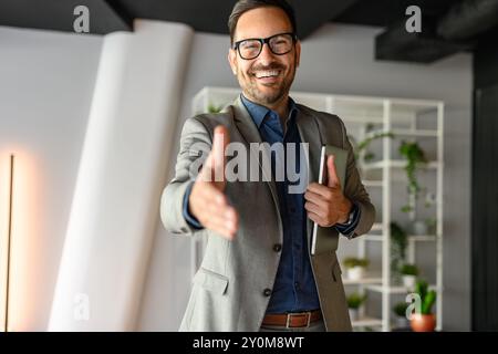 Porträt eines selbstbewussten, glücklichen Geschäftsmannes mit Brille, der Laptop hält und im Büro Handshake anbietet Stockfoto