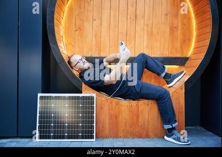 Bärtiger Mann mit Smartphone, das mit Photovoltaik-Sonnenkollektor verbunden ist. Integration nachhaltiger erneuerbarer Energien in den Alltag, demonstrieren die praktische Nutzung von Solarstrom für Ladegeräte. Stockfoto