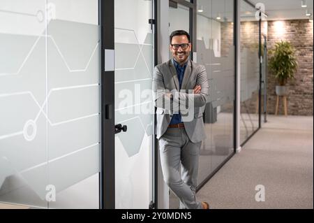 Junger glücklicher Geschäftsmann mit überkreuzten Armen, der sich an eine Glastür im modernen Büro lehnt und in die Kamera blickt Stockfoto