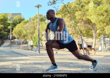 Afrikanischer Sportmann mit ärmellosem blauem Hemd, schwarzen Shorts dehnen sich die Beine oder wärmen sich im öffentlichen Park an sonnigen Sommertagen auf. Sport, h Stockfoto