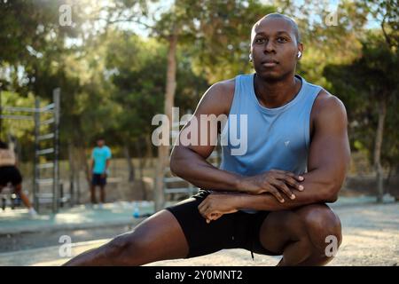 Afrikanischer Sportmann mit ärmellosem blauem Hemd, schwarzen Shorts dehnen sich die Beine oder wärmen sich im öffentlichen Park an sonnigen Sommertagen auf. Sport, h Stockfoto