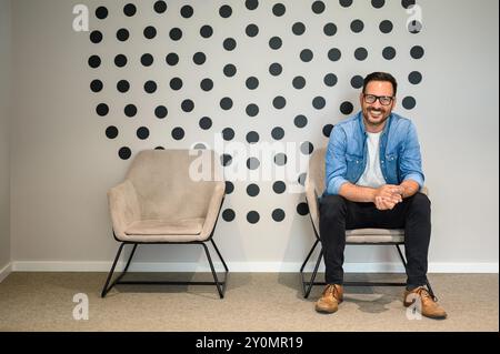 Porträt eines gutaussehenden, lächelnden Freelancers in Brille und mit gefesselten Händen auf Bürostuhl sitzend Stockfoto