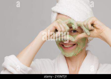 Eine Frau mit einem Handtuch auf dem Kopf lächelt, während sie eine grüne Gesichtsmaske aufträgt. Sie hat einen fröhlichen Ausdruck im Gesicht, der ihre Hautpflegeroutine zeigt Stockfoto