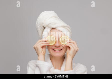 Ein glückliches Mädchen in Handtuch und Bademantel hält zwei Zitronenscheiben in der Hand und bedeckt ihre Augen mit einer Orange. Hautpflege und Feuchtigkeitskonzept Stockfoto