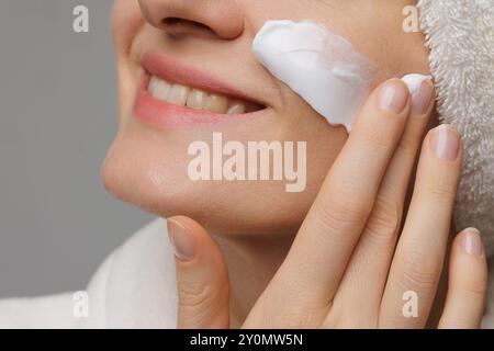 Eine Frau, die Creme auf ihr Gesicht aufträgt und sanft lächelt. Sie hat ein Handtuch um den Kopf gewickelt und ist in einer entspannten Umgebung, was auf eine Hautpflegeroutine hindeutet Stockfoto