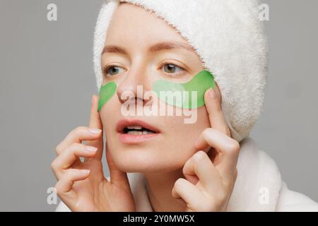 Eine Frau mit einem Handtuch auf dem Kopf und einem Bademantel lächelt, während sie grüne Gelflecken unter ihren Augen aufträgt Stockfoto
