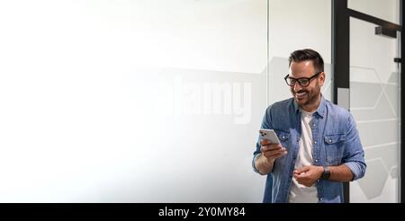 Gutaussehender Manager, der E-Mails über das Mobiltelefon liest, während er in einem modernen Büro an der Glaswand steht Stockfoto