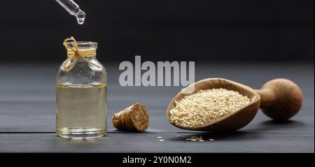 Glasflasche mit Sesamöl und rohen Sesamsamen in Holzschaufel auf schwarzem Holztisch. Öl tropft Stockfoto