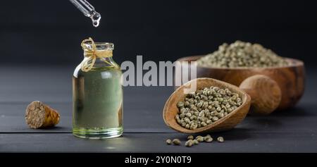 Haufen getrockneter Bio-Hanfsamen oder Hanfpflanzensamen in Löffel und Schüssel mit einem Glas Hanfsamenöl auf hölzernem Hintergrund, Öl tropft Stockfoto