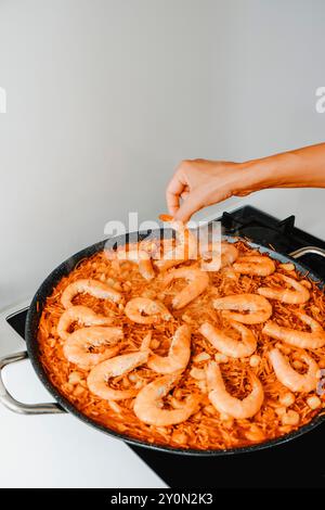 Ein Mann zeigt ein paar Garnelen zu einer spanischen Fideua, einer Nudelpaella, die in einer Paella-Pfanne gekocht wird, im Herd einer Hausküche Stockfoto