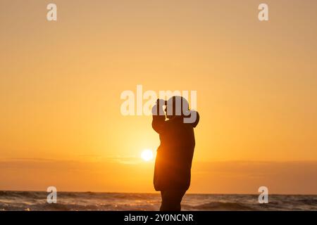 Silhouette einer isolierten Frau in Sportmütze, die an einem britischen Strand steht und bei Sonnenuntergang auf das Meer blickt. Stockfoto
