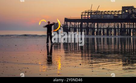 Man spinnt Feuer poi am Strand von Hastings, vor den verbrannten Überresten des alten Piers, Großbritannien. Stockfoto