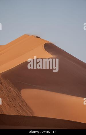 Wind weht Sand über die Spitze einer riesigen Sanddüne bei Sonnenaufgang in Sossusvlei, Namibia Stockfoto