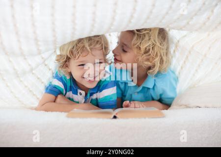 Lesebuch für Kinder im Bett unter der Strickdecke. Zwei Brüder spielen zusammen. Kinder gemütliches Zimmer im Hyggge-Stil. Kleiner Junge, der Hausaufgaben macht, bevor er schläft. Stockfoto