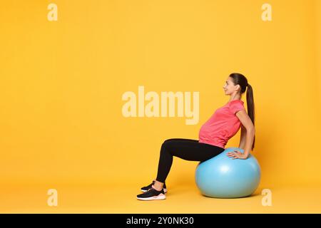 Schöne schwangere Frau, die Übungen auf dem Fitball vor gelbem Hintergrund macht Stockfoto