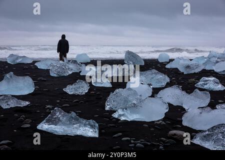 Diamond Beach in Island - transparente Eisberge, die an Land gewaschen werden, auf schwarzem Sandstrand mit der Silhouette einer Person, die am Strand steht, am Meer. Stockfoto