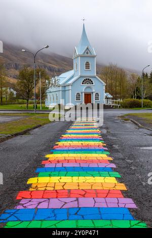 Seydisfjordur, Island, 19.05.2022. Blaue Kirche Seydisfjordur (Seyðisfjarðarkirkja) mit einer Straße mit Regenbogenpflaster, nebelige Berge im Hintergrund Stockfoto