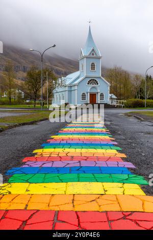 Seydisfjordur, Island, 19.05.2022. Blaue Kirche Seydisfjordur (Seyðisfjarðarkirkja) mit einer Straße mit Regenbogenpflaster, nebelige Berge im Hintergrund Stockfoto