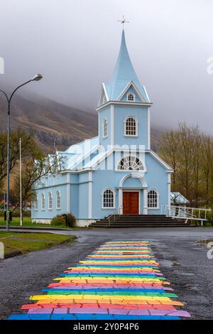 Blaue Kirche Seydisfjordur (Seyðisfjarðarkirkja) mit einer Straße mit Regenbogenpflaster, nebelige Berge im Hintergrund, Island. Stockfoto