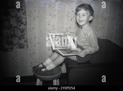 1950er Jahre, historisch, ein kleiner Junge, Hemd und Bowtie, ein großes Lächeln im Gesicht, auf einem Schrank sitzend, Füße auf einem Hocker und mit einem Tier-/Dschungelbuch auf dem Schoß. Stockfoto