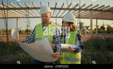 Zwei kaukasische Arbeiter Uniform Hardhat weibliches Mädchen männlicher Mann Mann Ingenieure Partner halten Blaupausen ökologischem Erbauer zeigt Baustellenspezialisten Stockfoto