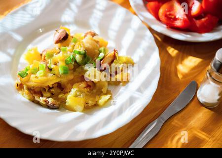 Gebackene Kartoffel mit Wurst auf weißem Teller, Besteck, Salzstreuer und frischer Tomatensalat auf Holztisch in Streifenbeleuchtung, Nahaufnahme. Stockfoto