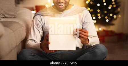 Überraschter Afro-Mann, der ein Geschenk gegen den Weihnachtsbaum eröffnet Stockfoto