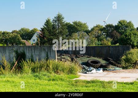 In einem verlassenen Industriegebiet befinden sich ausrangierte Reifen und verstreute Trümmer, umgeben von hohem Gras und Bäumen. Häuser und eine Windturbine sichtbar in t Stockfoto