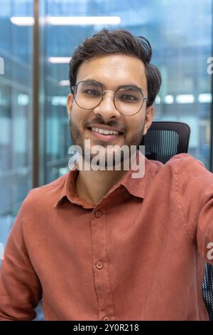 Junger Mann mit Bart und Brille, der im Büro Videogespräche führt. Er lächelt warm, während er direkt in die Kamera schaut und Bilder mit dem Telefon aufnimmt. Professionelle Kleidung deutet auf den Bürokontext hin. Stockfoto