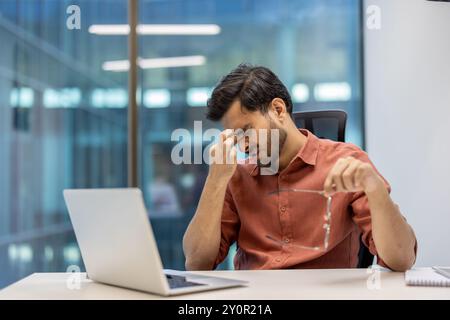 Büroangestellter, der am Schreibtisch sitzt, hat Stress und Kopfschmerzen, während er eine Brille hält. Das Bild erfasst Ermüdungs- und Druckgefühle. Begriff von Arbeitsstress und psychischer Gesundheit. Stockfoto