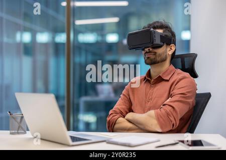 Mann im Büro, der ein Virtual-Reality-Headset trägt und die immersive Technologie erforscht. Am Schreibtisch mit Laptop sitzen. Das Konzept umfasst Technologie, Innovation und ein futuristisches Arbeitsumfeld. Stockfoto