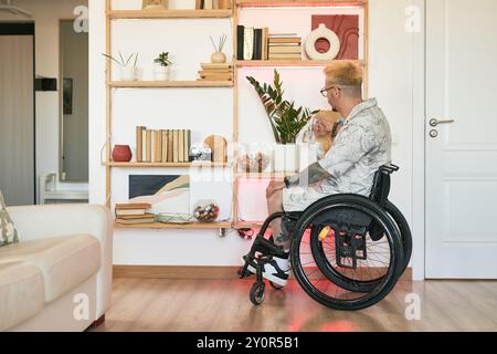 Mann mit Tätowierungen im Rollstuhl, der neben einem Bücherregal mit dekorativen Gegenständen sitzt, in einem modernen Wohnzimmer mit Holzboden und weißen Wänden, seitlich gesehen Stockfoto