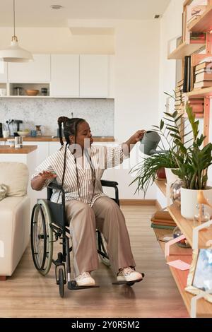 Afroamerikanische Frau im Rollstuhl gießt Wasser auf Pflanzen in moderner Küche mit Holzregalen und gemütlichem Dekor Stockfoto