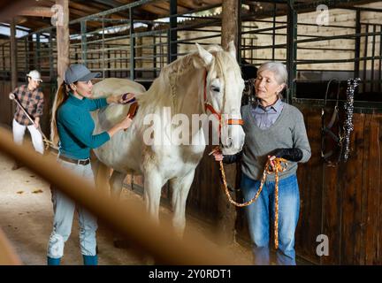 Arbeiterinnen bürsten nach dem Reiten im Stall weißes Rennpferd Stockfoto