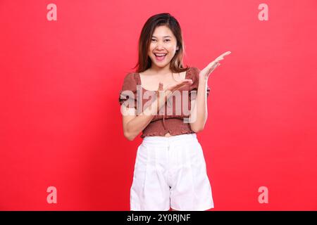 Fröhliche asiatische Frau, beide Handflächen zeigen nach links, tragen eine braune Bluse und Shorts auf rotem Hintergrund. Für Mode, Lifestyle und Stockfoto