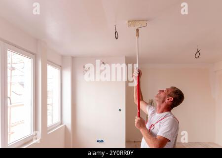 Bauarbeiter Maler Mann mit Malerwalze in der Hand und Eimern mit Produkten, um die Wand und die Decke im Gebäude zu restaurieren und zu lackieren Stockfoto