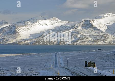 Schneebedeckte Straße, die zu einem See führt, umgeben von Bergen, mit einem Auto am Rande der Winterlandschaft, Skagarfjoerdur, Nordisland, Island, Europa Stockfoto