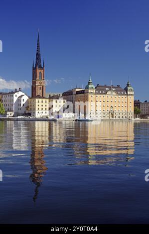 Malerischer Blick auf einen Kirchturm und historische Gebäude entlang eines ruhigen Ufers mit klarem Wasser unter einem sonnigen Himmel, Gamla Stan, Altstadt, Stoc Stockfoto