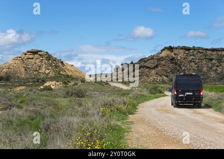 Schwarzer Van auf einer Schotterstraße in einer hügeligen, grünen Landschaft unter blauem Himmel mit Wolken, Wohnmobil, Wohnmobil, Bardenas Reales Naturpark, Wüste, Halbdörfer Stockfoto