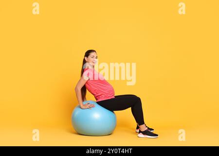 Schöne schwangere Frau, die Übungen auf dem Fitball vor gelbem Hintergrund macht Stockfoto