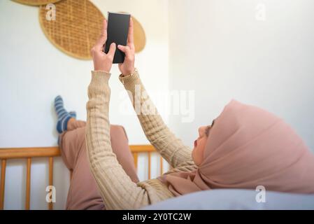 Eine arabische muslimische Frau liegt auf einem Bett und schaut auf ihr Handy. Sie trägt einen Hijab und hat einen rosa Schal um den Kopf Stockfoto