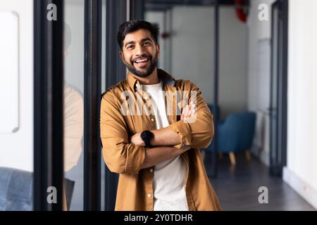 Lächelnder indischer Mann in lässiger Kleidung, der mit überkreuzten Armen im modernen Büro steht Stockfoto